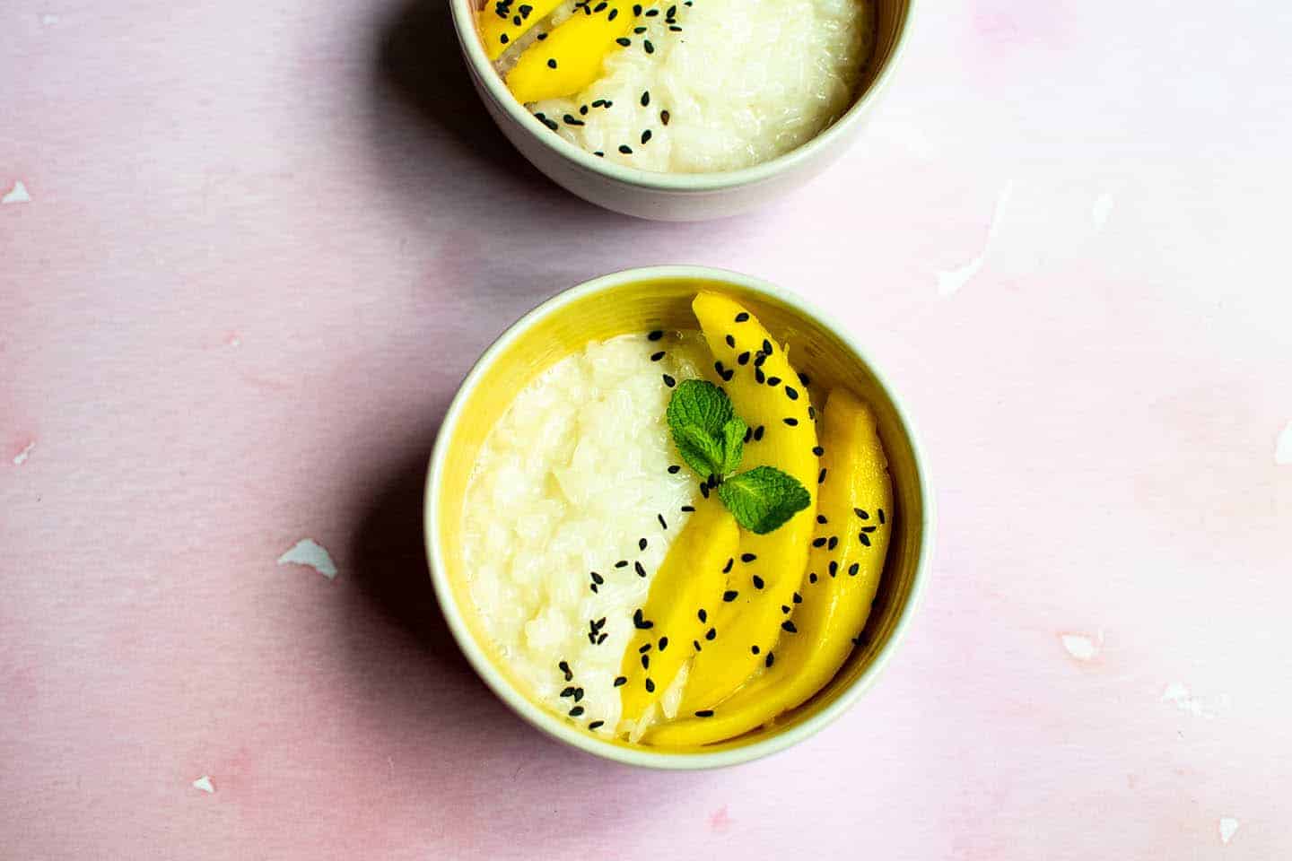 A closer photo of one of the bowls of mango sticky rice. It's on a pink worktop and has black sesame seeds and fresh mint scattered on top.