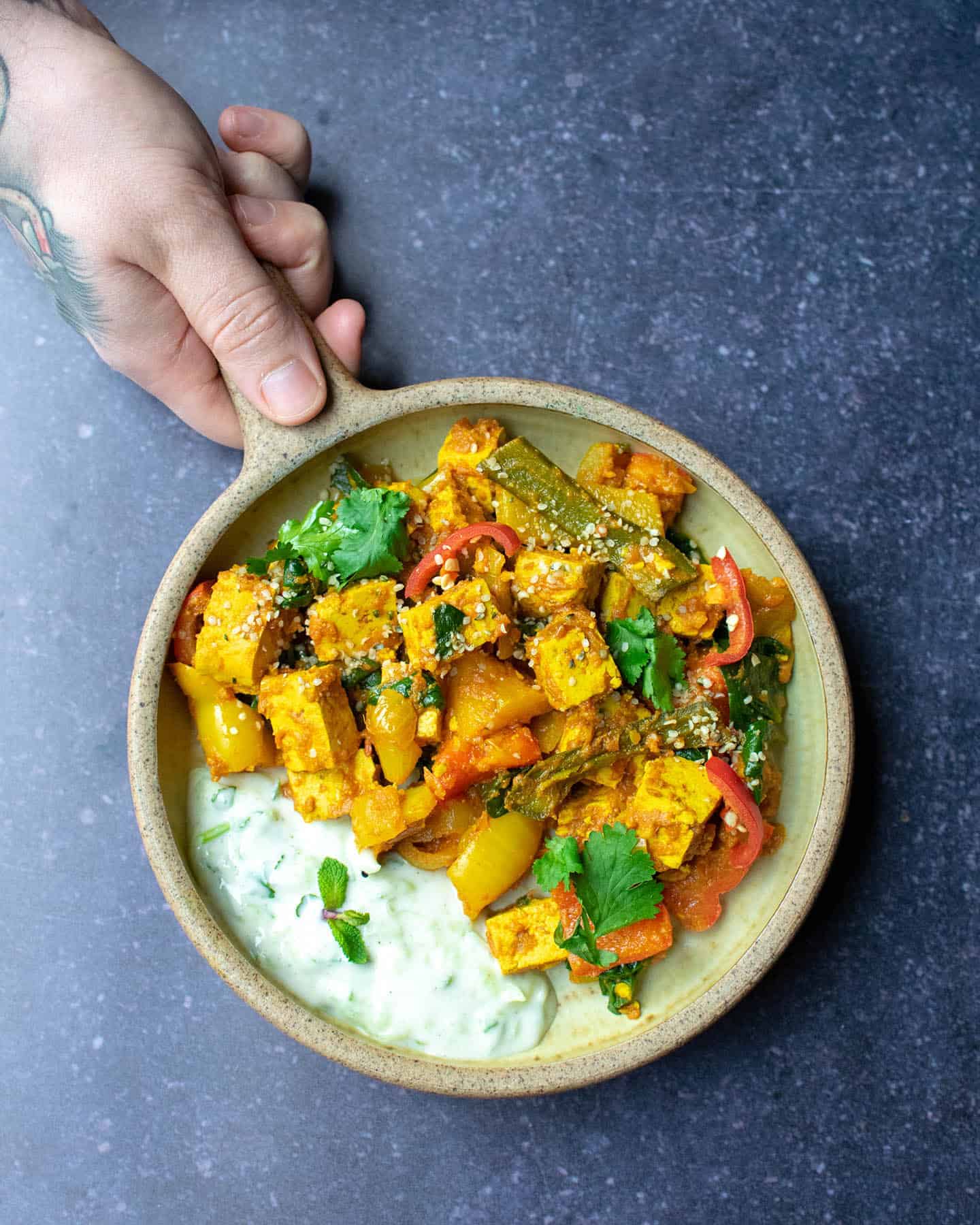 A bowl of vegetable bhuna with a man's hand holding the handle of the dish