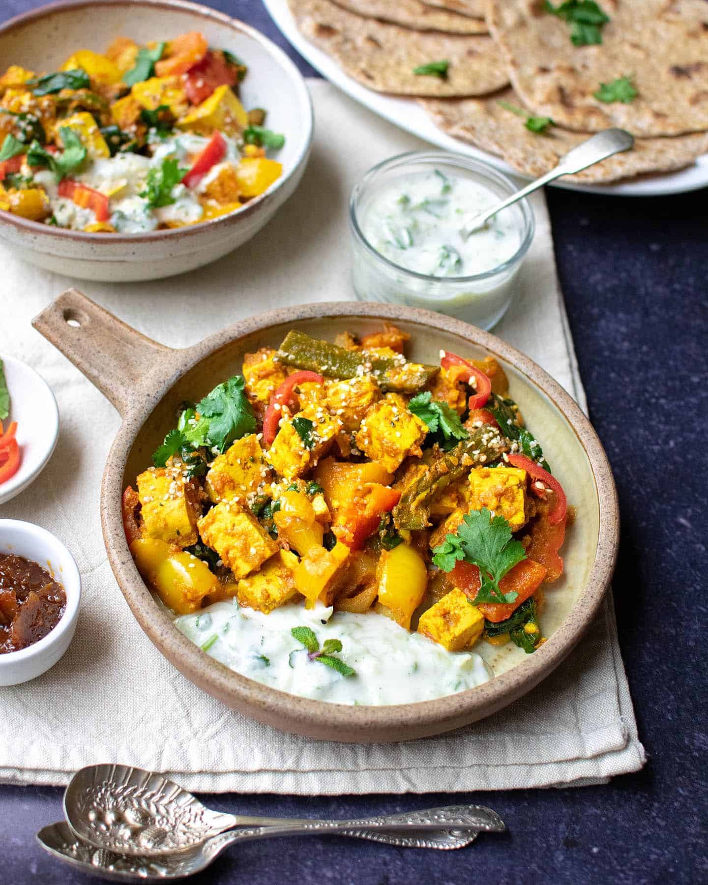 Two bowls of veg curry with bread and other condiments in the background