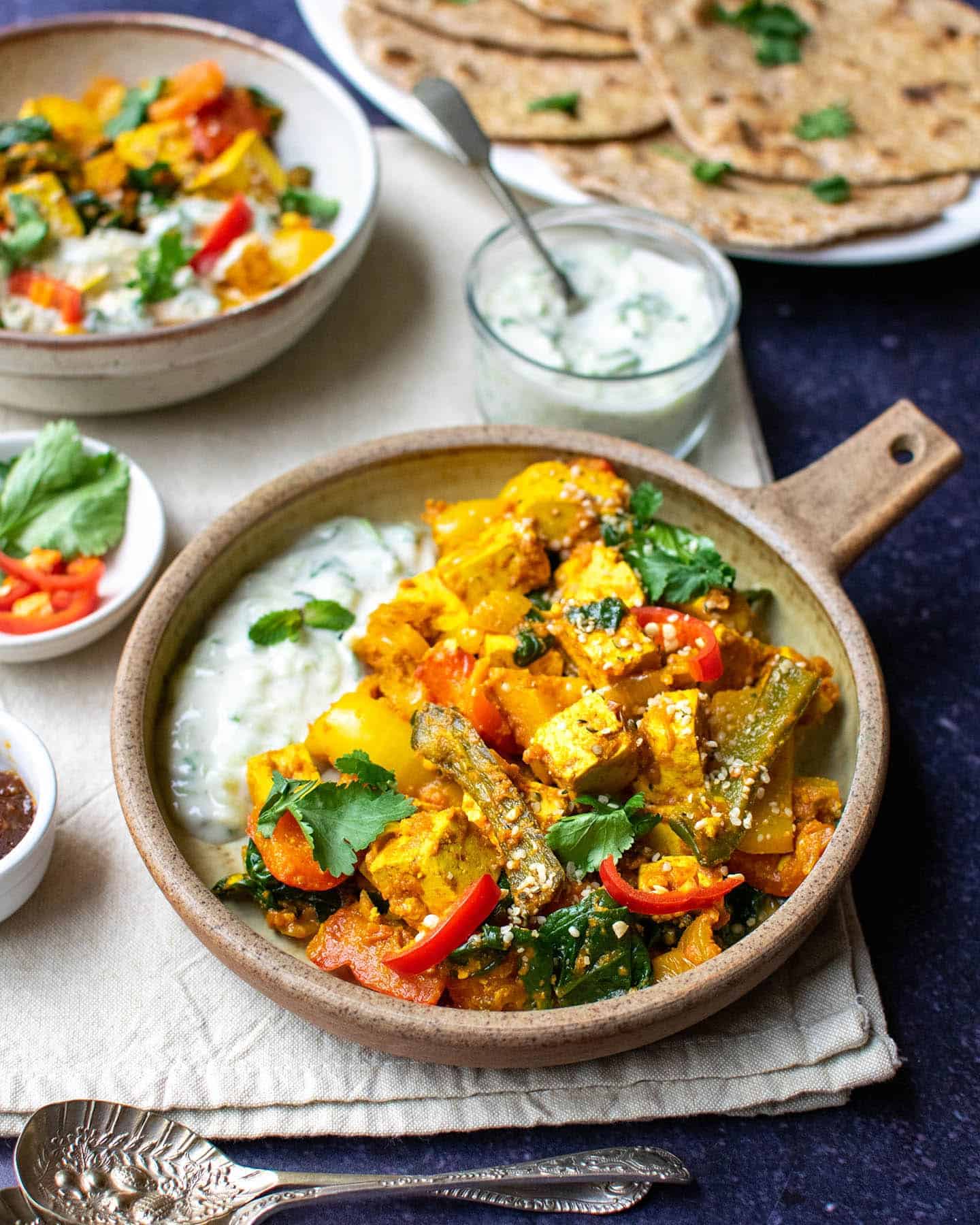 Vegetable bhuna in a round dish with a handle, yoghurt behind it and another bowl of curry in the background