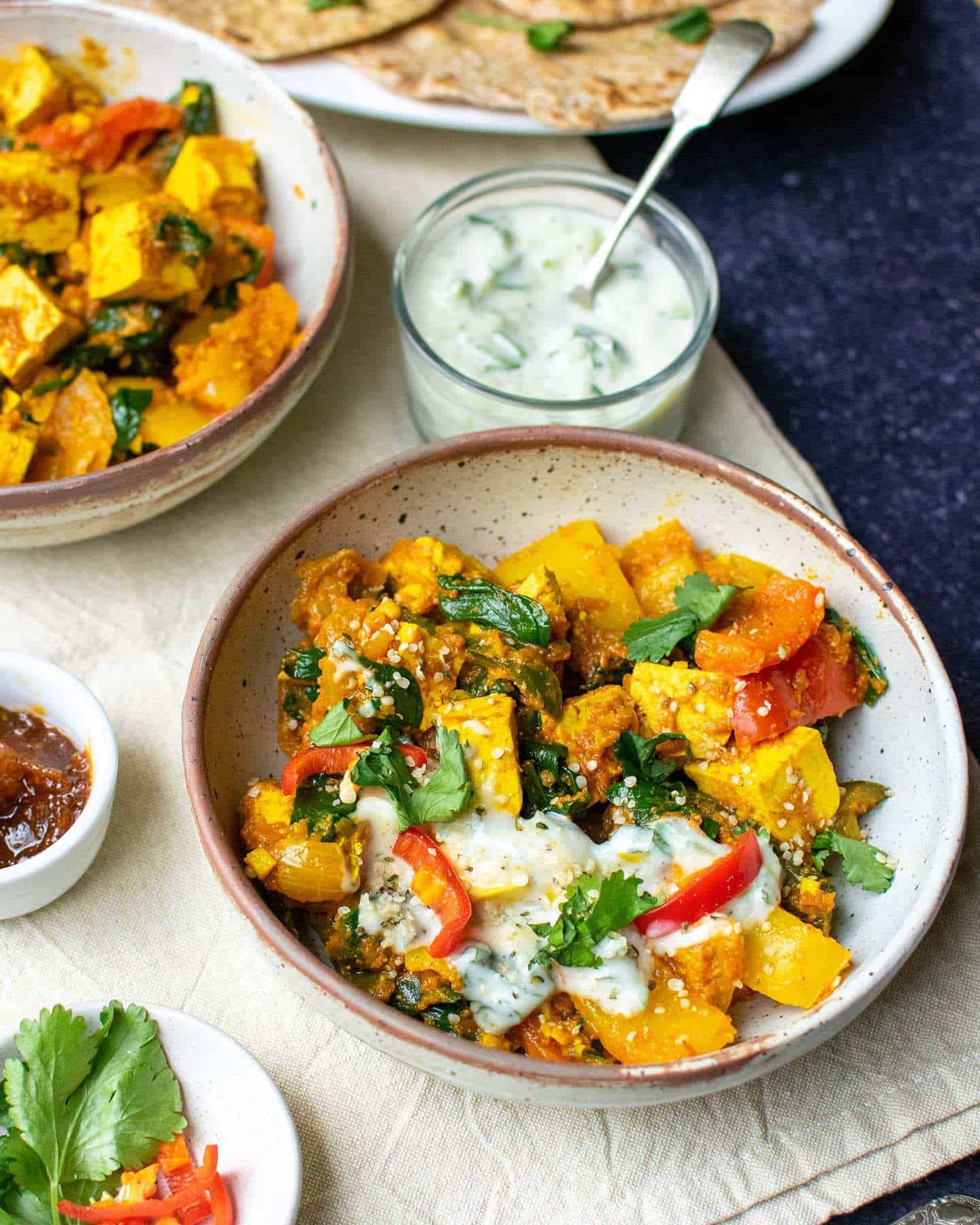 A bowl of vegetable curry with tofu in a bowl, with yoghurt and roti in the background and a dish of coriander and chillies and a small pot of chutney