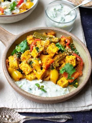 Vegetable bhuna with peppers, spinach and tofu in a round dish with a handle, with spoons and yoghurt and chutney around it