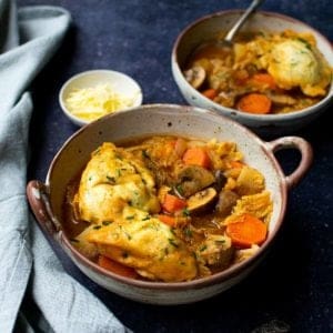 A bowl with handles full with vegan stew and dumplings, a pot of vegan vegan in the background along with another smaller bowl of stew