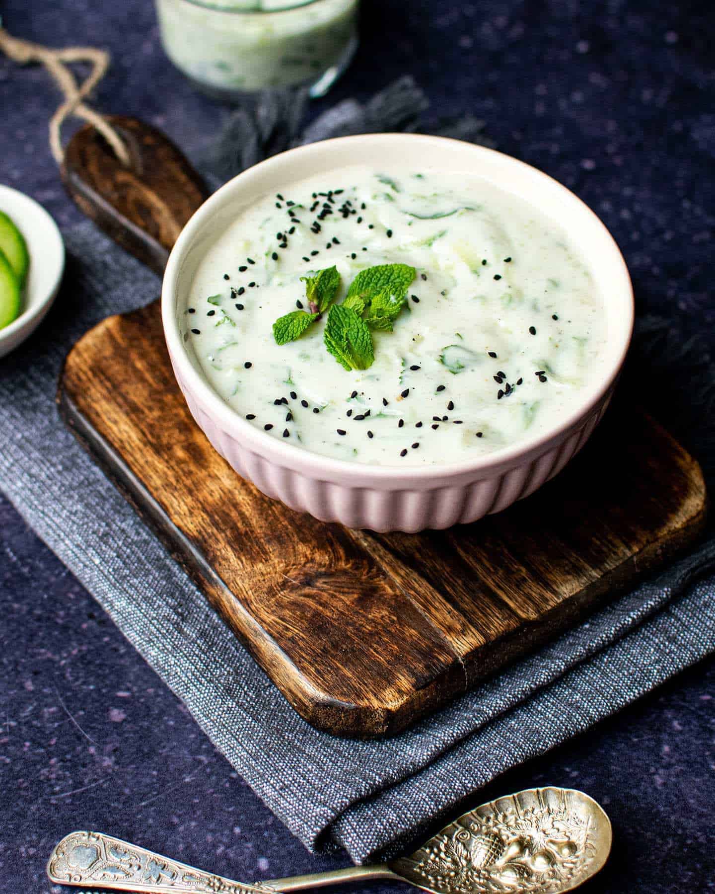 A pink bowl of vegan cucumber raita with mint on top and two spoons in the foreground