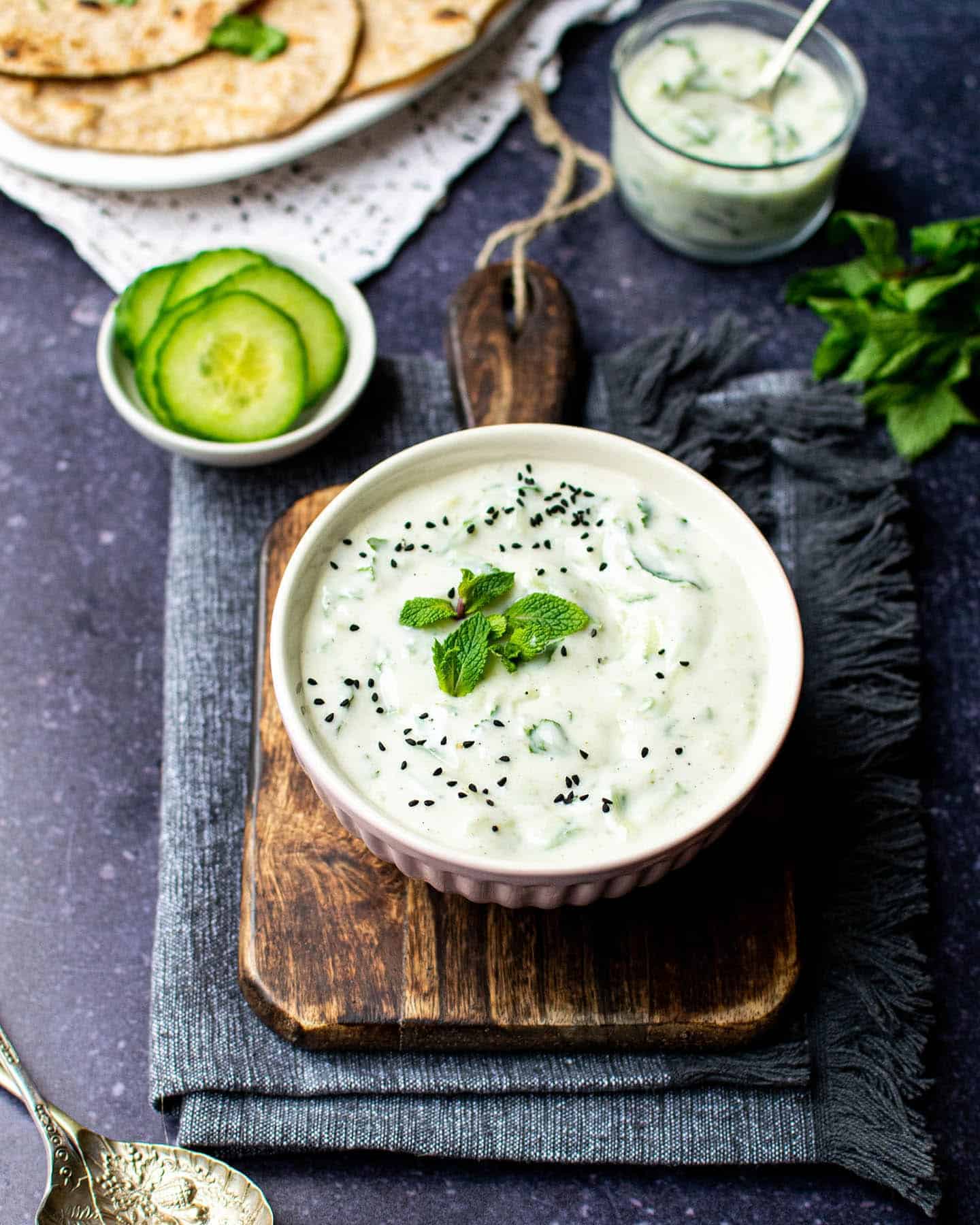 A top down view of a bowl of vegan cucumber raita with a pot of cucumber, roti in the background and fresh mint