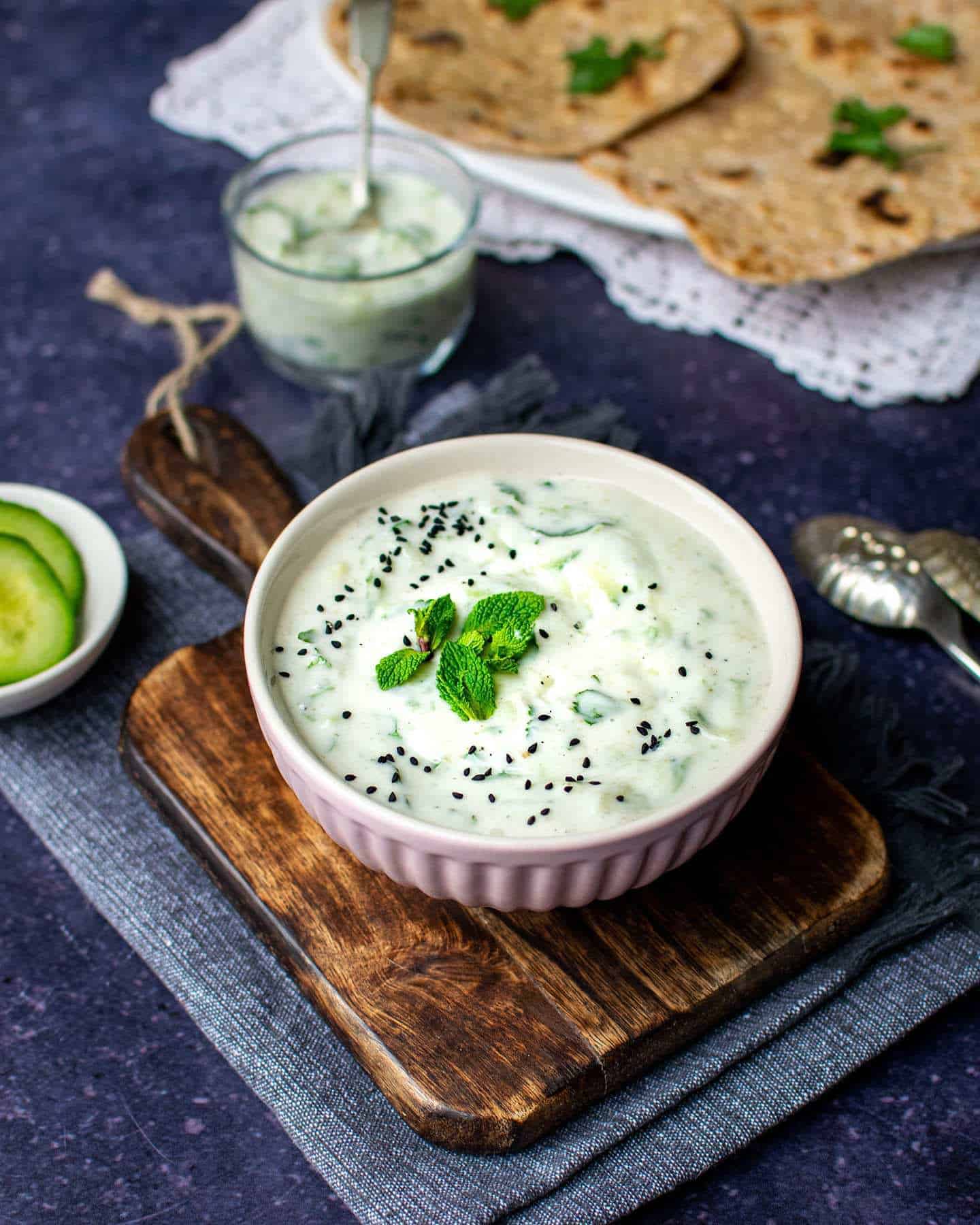 A bowl of raita with fresh mint and nigella seeds sat on top of a chopping board, with cucumber in a small pot to the left, with roti to the back of the image on a plate with coriander on top