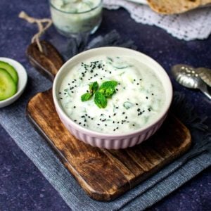 A pink bowl of vegan raita with nigella seeds and mint leaves on top, on a chopping board with a second glass of yoghurt in the background