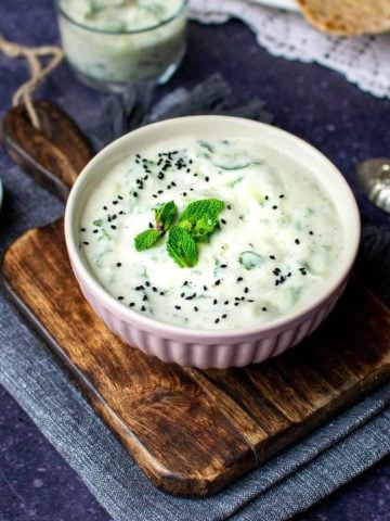 A pink bowl of vegan raita with nigella seeds and mint leaves on top, on a chopping board with a second glass of yoghurt in the background