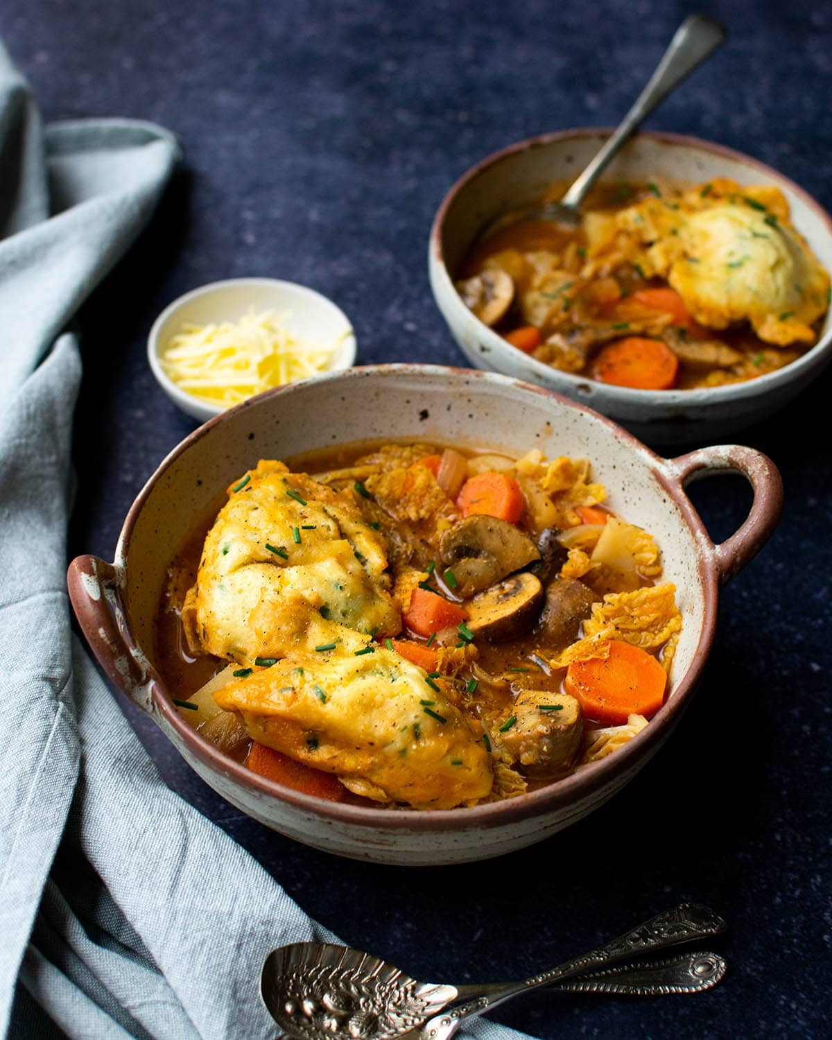 Two bowls of vegan stew topped with fluffy dumplings, vegan cheese in a small bowl.