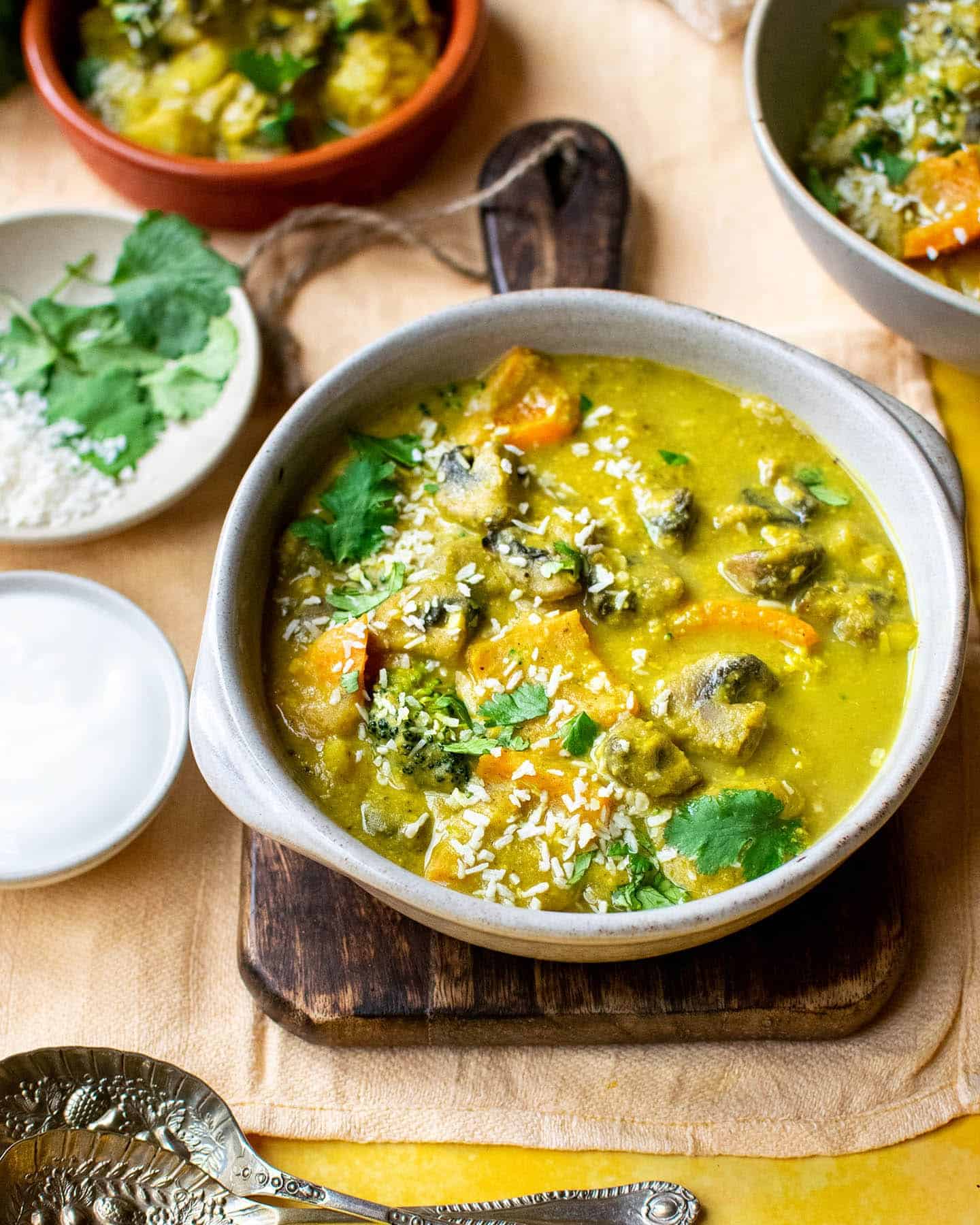 A bowl of vegan korma with spoons in front of it, vegan yoghurt fresh coriander and another 2 bowls of curry