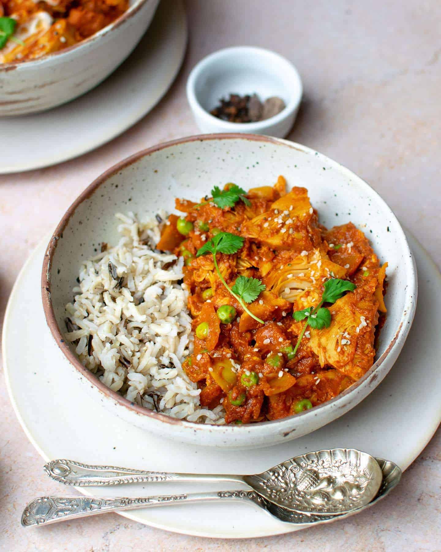 A bowl full of this recipe and rice with spoons in the foreground and spices in a small round white pot in the background