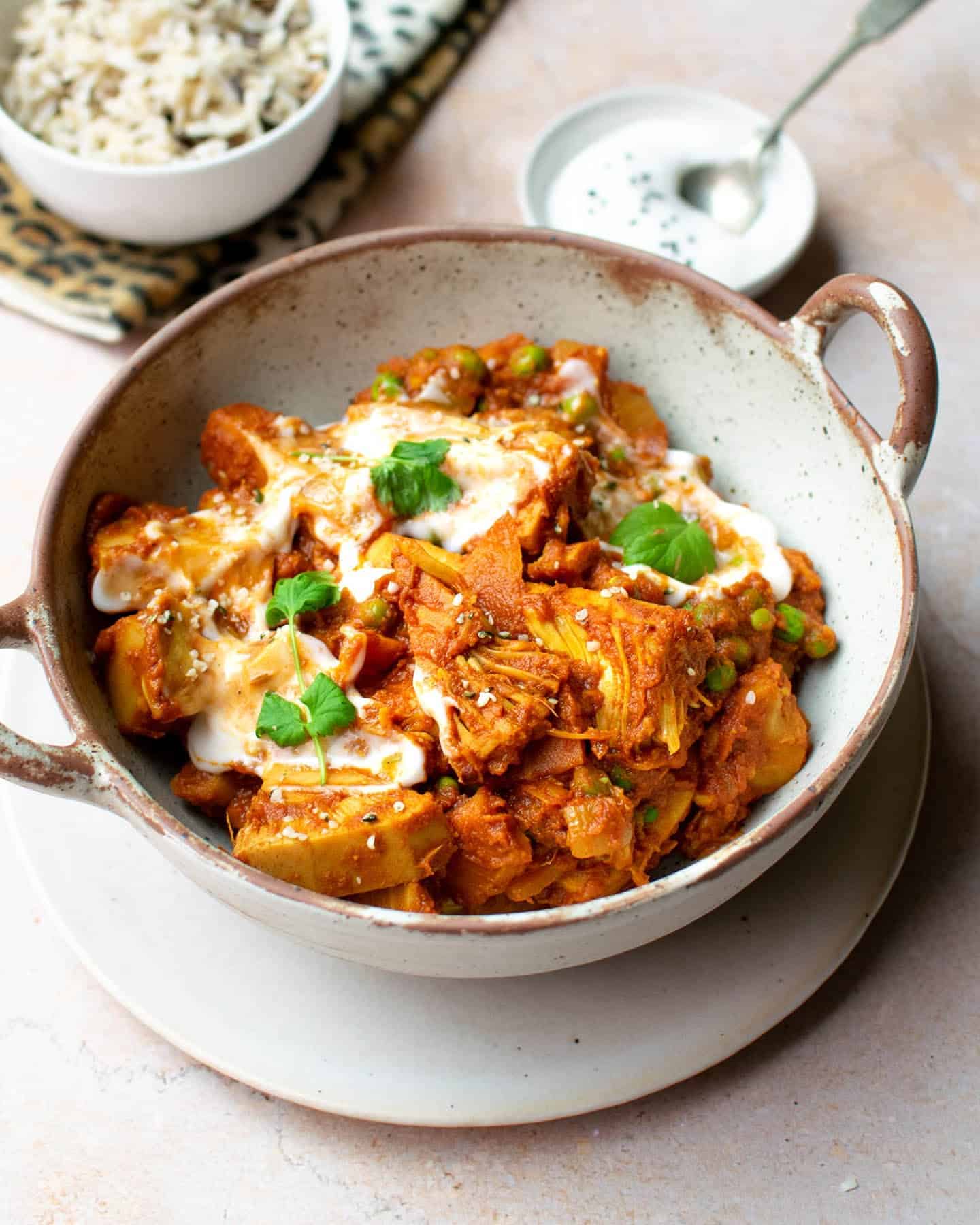 Portrait image of vegan jackfruit curry in a bowl with handles on either side, sitting on top of a plate