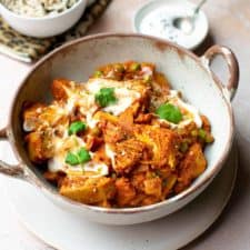 Jackfruit curry in a bowl with handles with vegan yoghurt and coriander on top. There's a small bowl of yoghurt in the background as well as a bigger bowl of rice