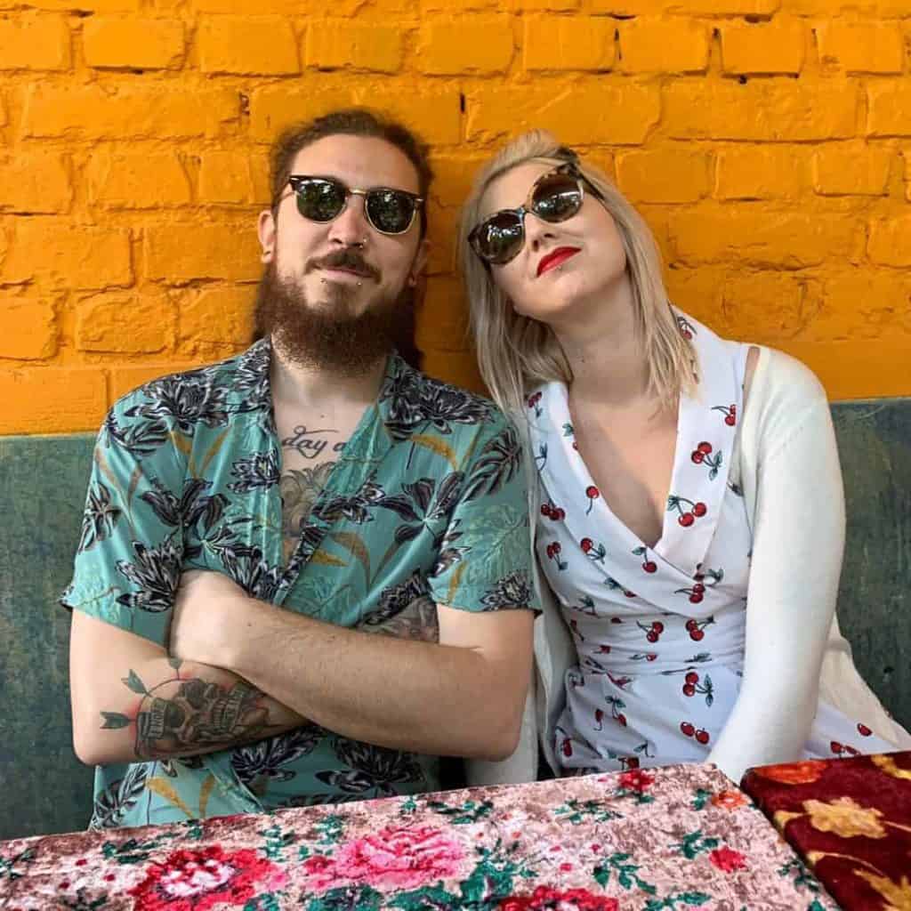 Jess and Dan wearing sunglasses, sat against a painted brick wall