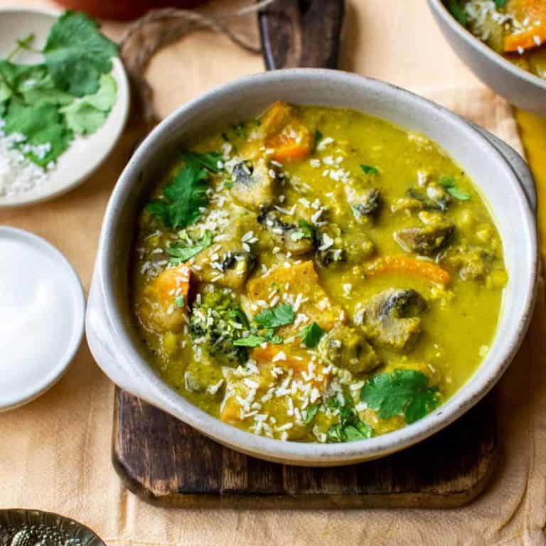 Mushroom Korma in a white bowl, on top of a small chopping board, with condiments all around it, including yoghurt, coriander and desiccated coconut flakes.