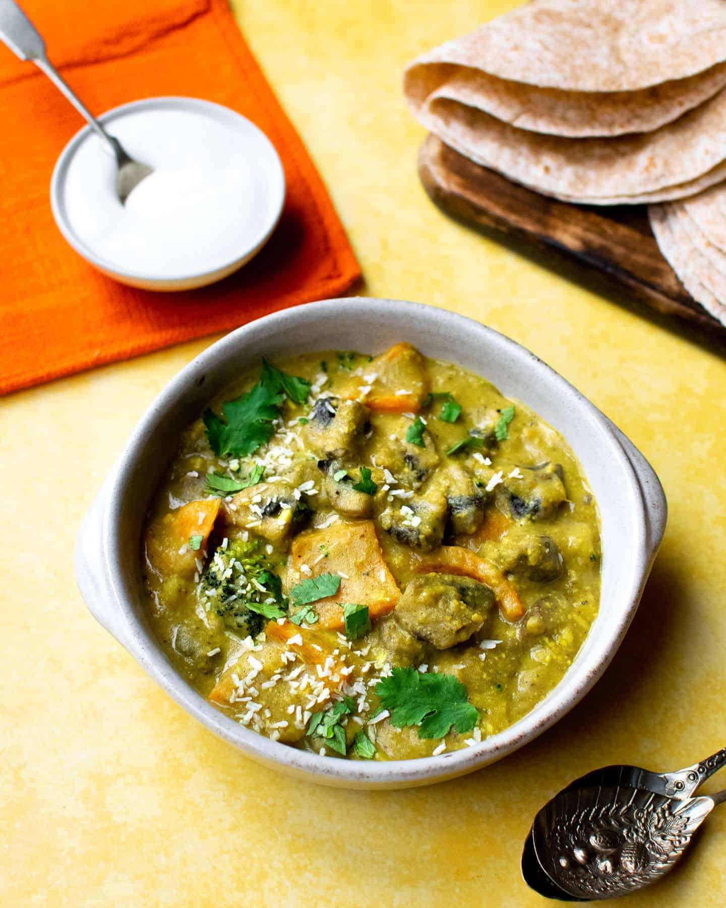 Curry in a bowl with spoons, parathas and vegan yoghurt sitting on an orange cloth in the background