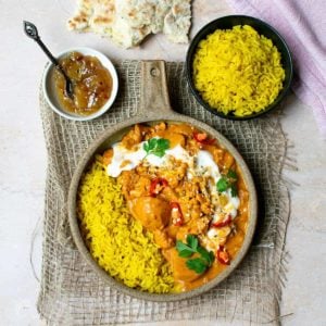 A bowl of vegetable tikka masala on hessian background with rice to one side of the bowl. There's chutney and naan in the background