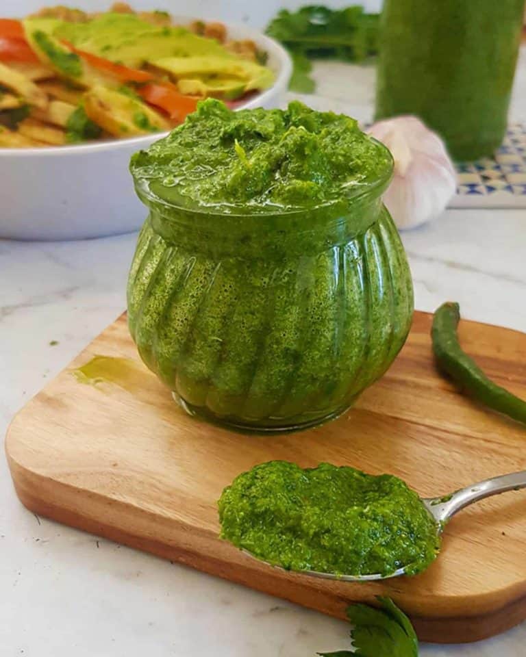 A glass jar of green chutney on a chopping board with a spoon full of chutney in the foreground, there's a dish of food in the background too