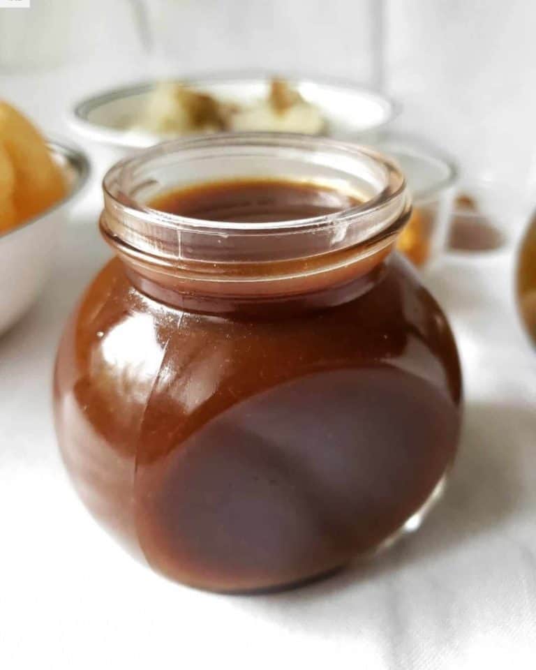 A jar of brown tamarind sauce full to the top with some bowls in the background but it's not clear what's in them