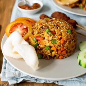 Square image showing vegan nasi goreng in a mound on a grey plate surrounded by cucumber, tomato and satay skewers
