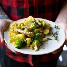 Dan's hands holding a white plate of vegan potato hash where you can see broccoli, swede mash, vegan chicken and fresh thyme, held up against a checked red shirt