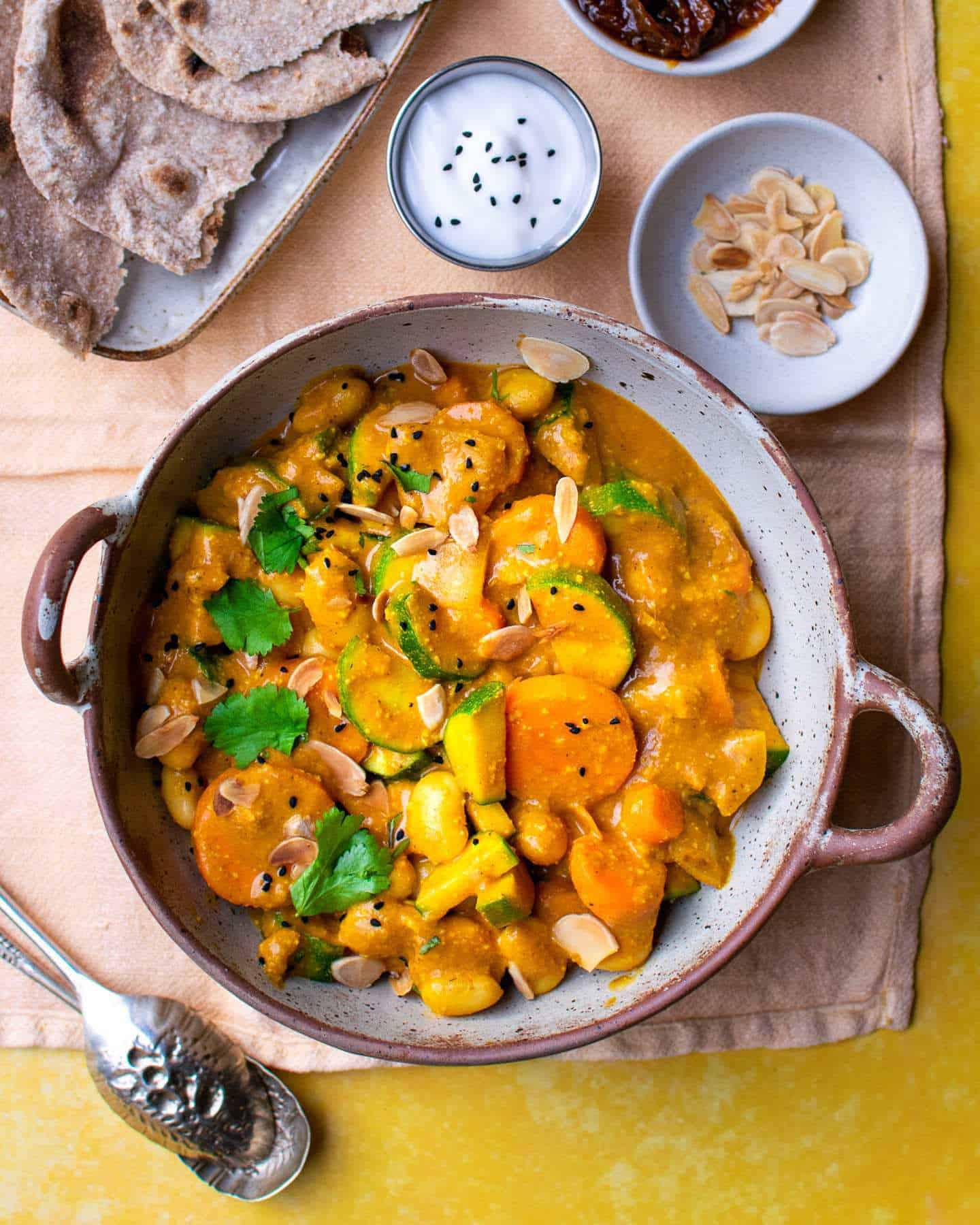 A vegan curry with butter beans, courgette and carrots in an orange creamy sauce, served in a big bowl with two handles and surrounded by yoghurt, flaked almonds and Indian bread