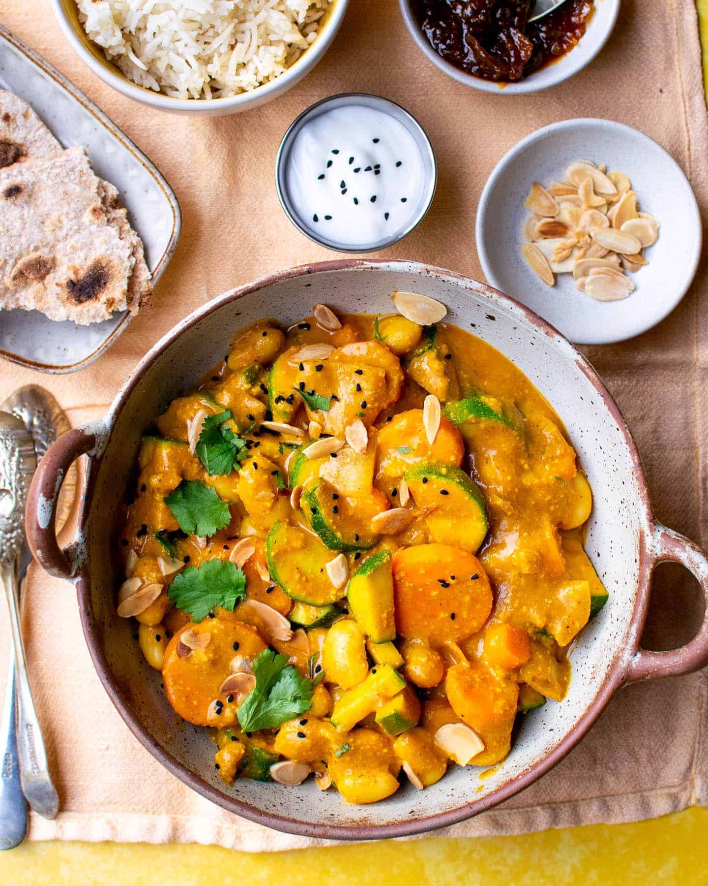 A big bowl of vegan passanda with serving suggestions around it, including toasted almond flakes, rice, roti and vegan yoghurt. All set on an orange background.