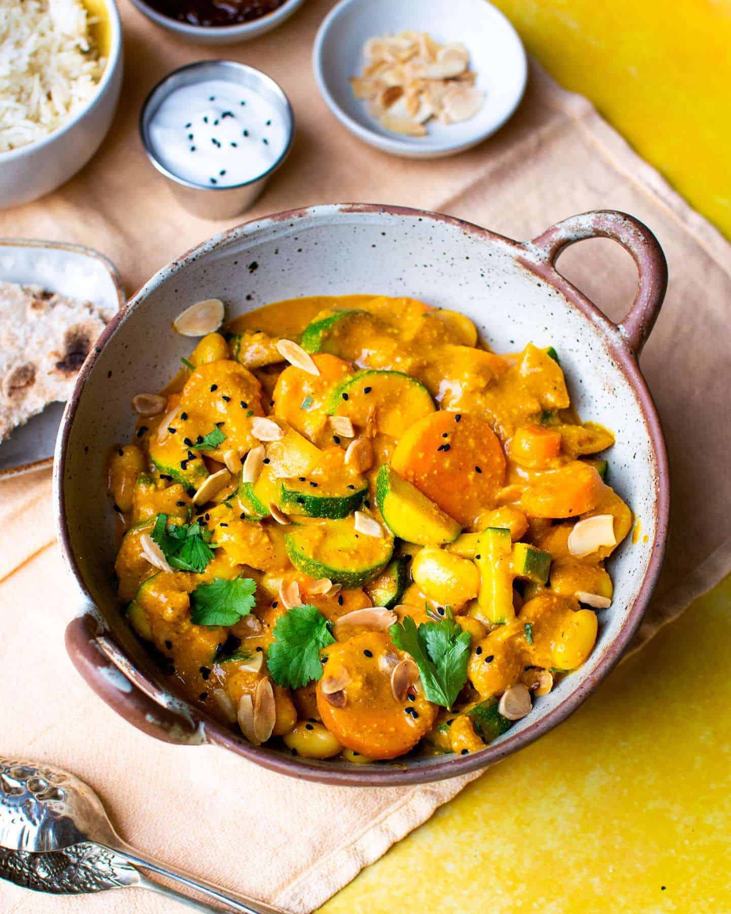 Vegetable passanda in a bowl featuring courgettes, carrots and butter beans on an orange and yellow background, with condiments around the bowl in small pots