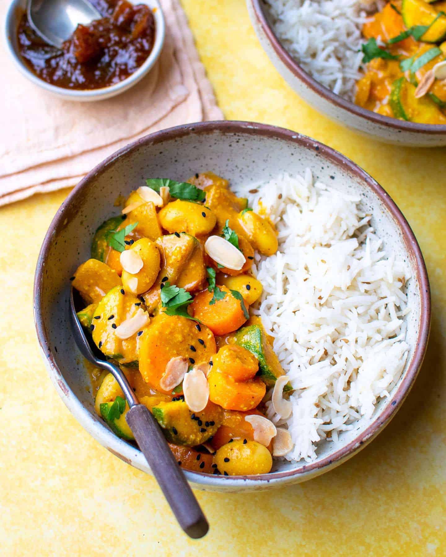 A small bowl of vegetable passanda and rice with a spoon with a wooden handle in it, ready to be eaten. There's a pot of chutney in the left hand corner and another bowl of curry just visible in the right hand corner