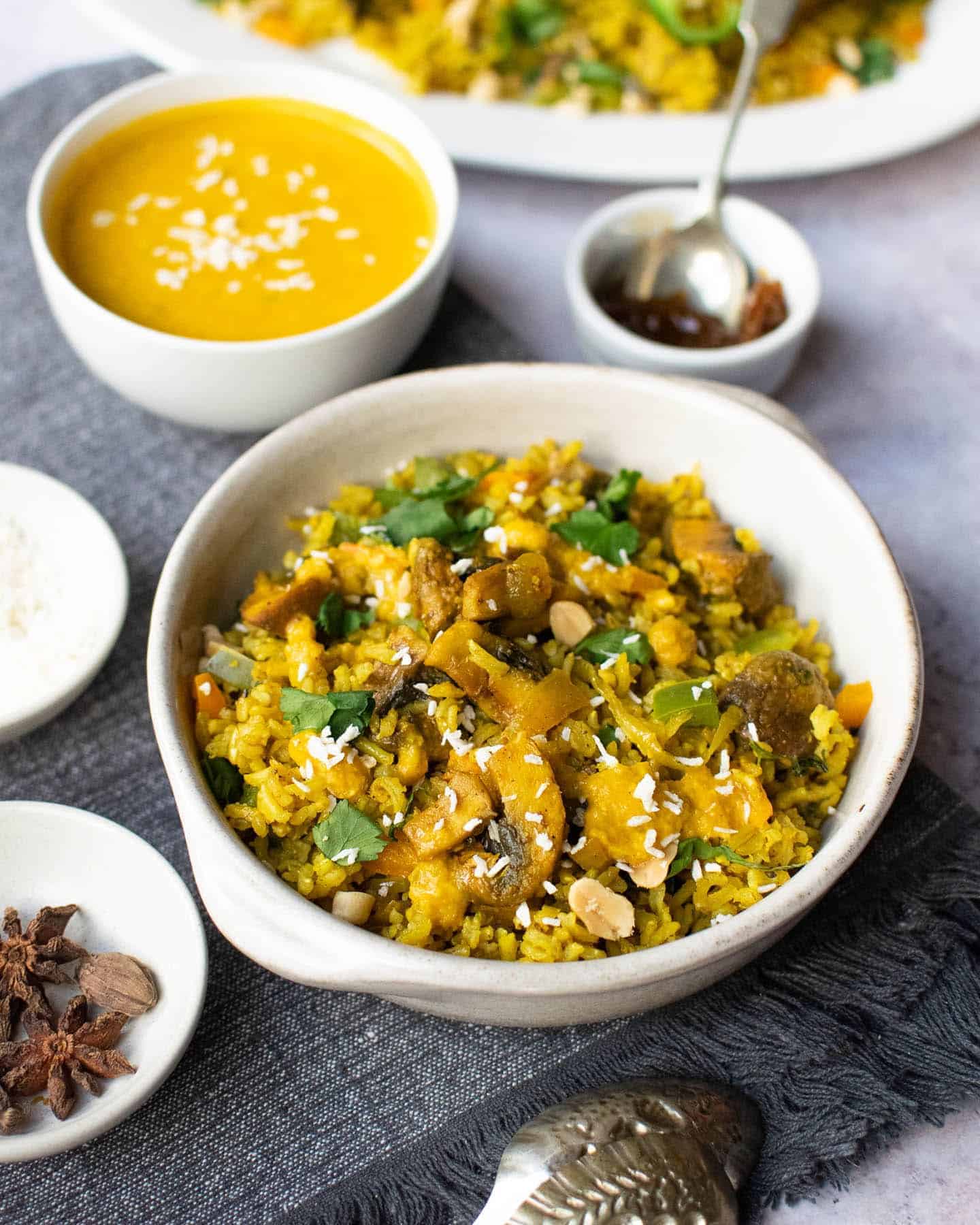 Mushroom biyani in a small white dish with two ridges for handles, with spoons next to it and condiments surrounding it too. They include orange biryani gravy, chutney and a big plate of rice in the background