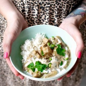 Hands holding up a green bowl filled with vegan korma showing potato, aubergine, fresh coriander and sauce. The background shows a leopard print dress