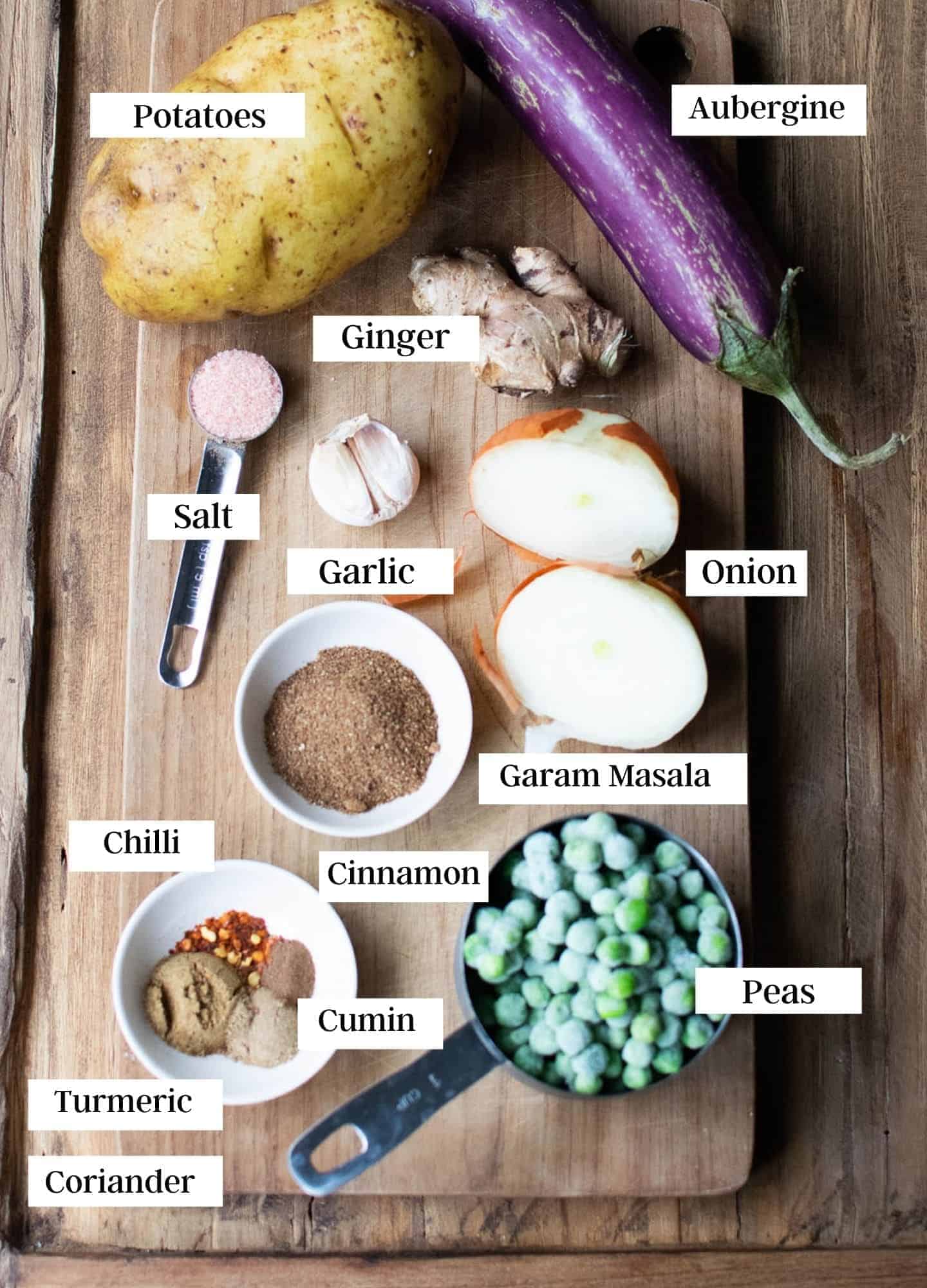Top down view of ingredients laid out on top of a chopping board. Showing aubergine, potato, peas, onion, garlic, ginger and powdered spices and salt