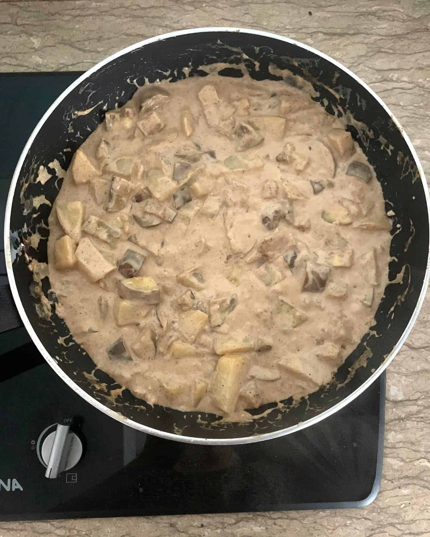 A light brown curry in a black pan with chunks of potatoes and aubergines visible in a thick and creamy brown sauce