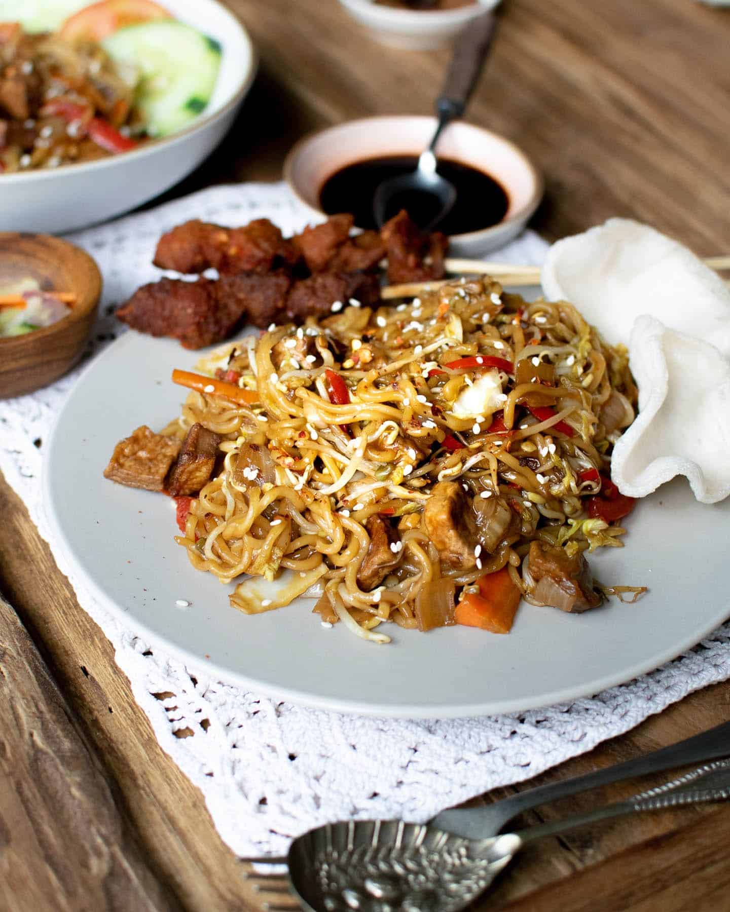 Vegan mi goreng on a grey plate with spoons in the foreground, soy sauce in the background and a plate of noodles to the left in the background