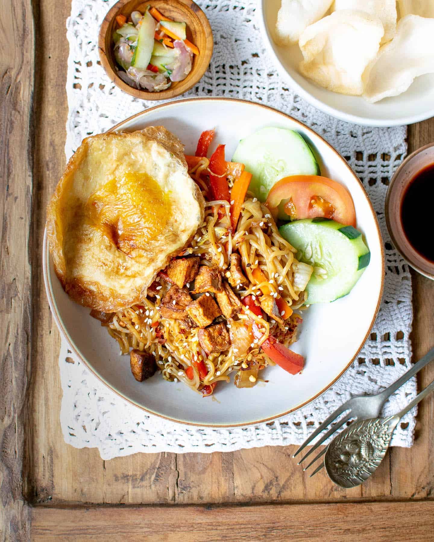 To down view of a plate of vegan mi goreng with a vegan fried egg to one side, cucumber and tomato and small bowls of condiments surrounding it