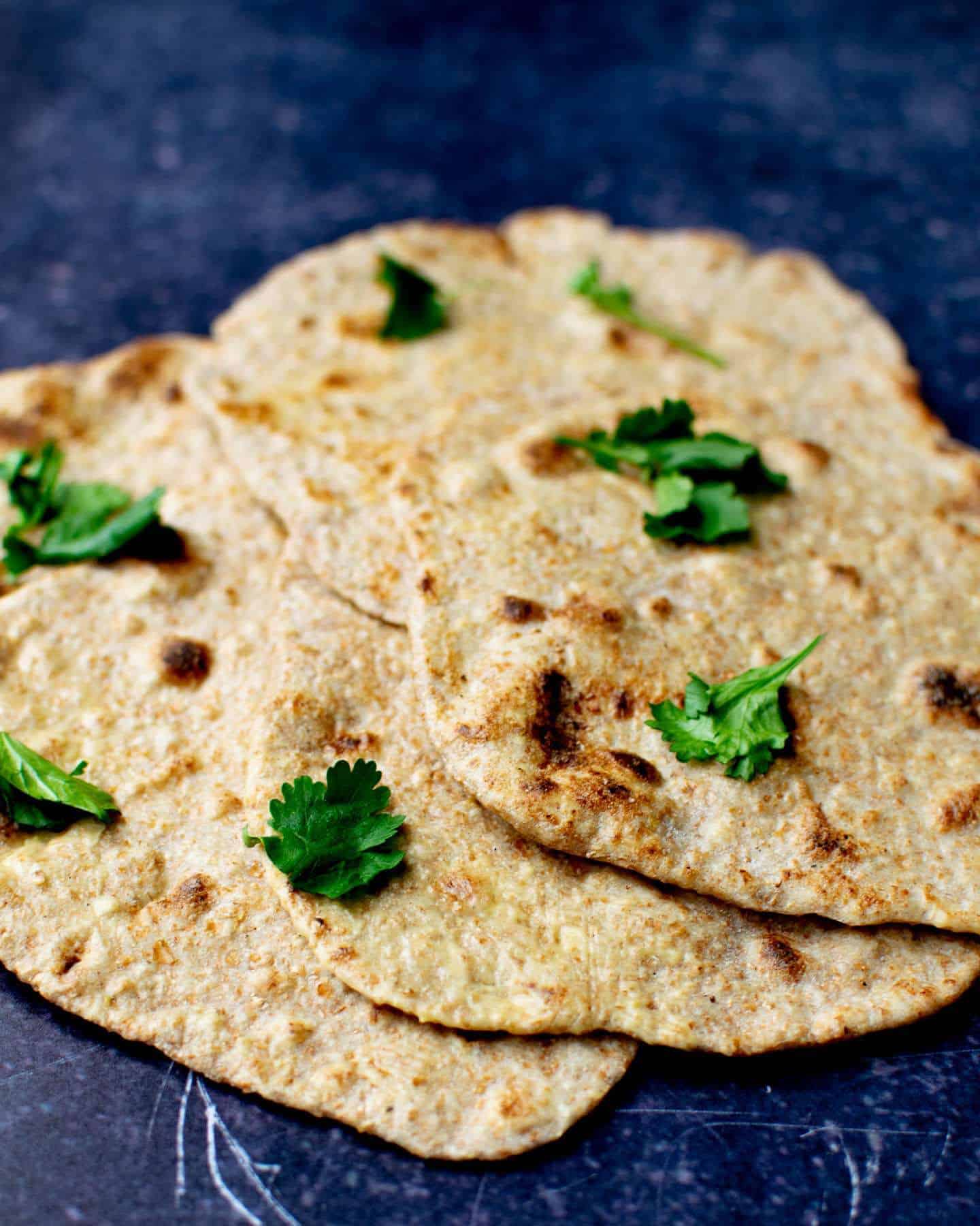 Close up portrait image of vegan roti laid out on top of each other with fresh coriander on top, all on a dark blue background