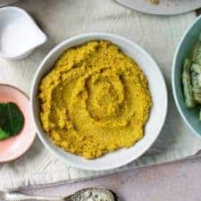 Square image, top down view of a white bowl full with Balinese spice paste with spoons, and other plates surrounding it