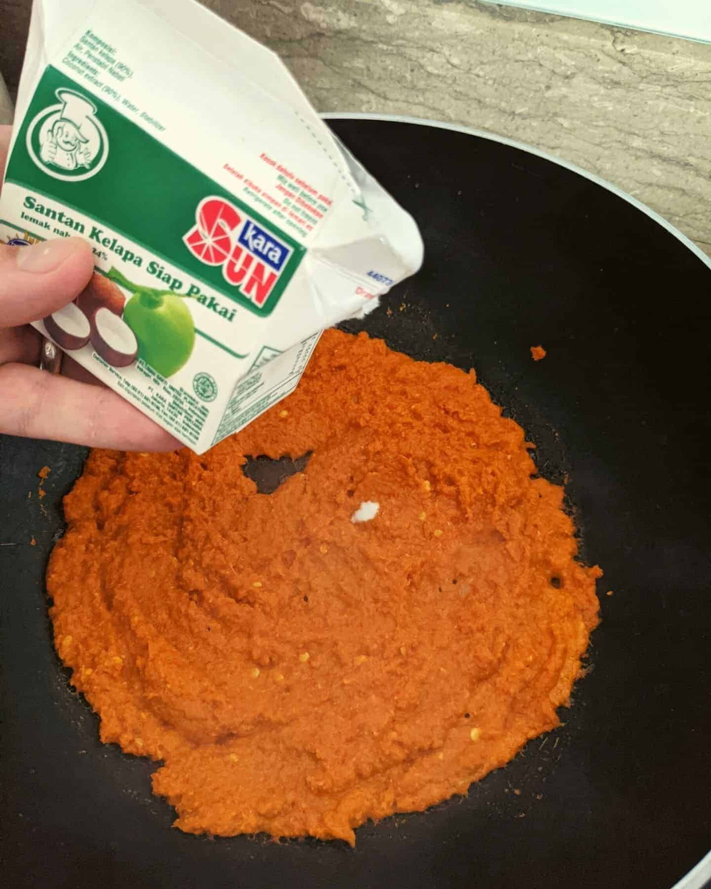 A carton of coconut milk being held over a wok with curry paste in it