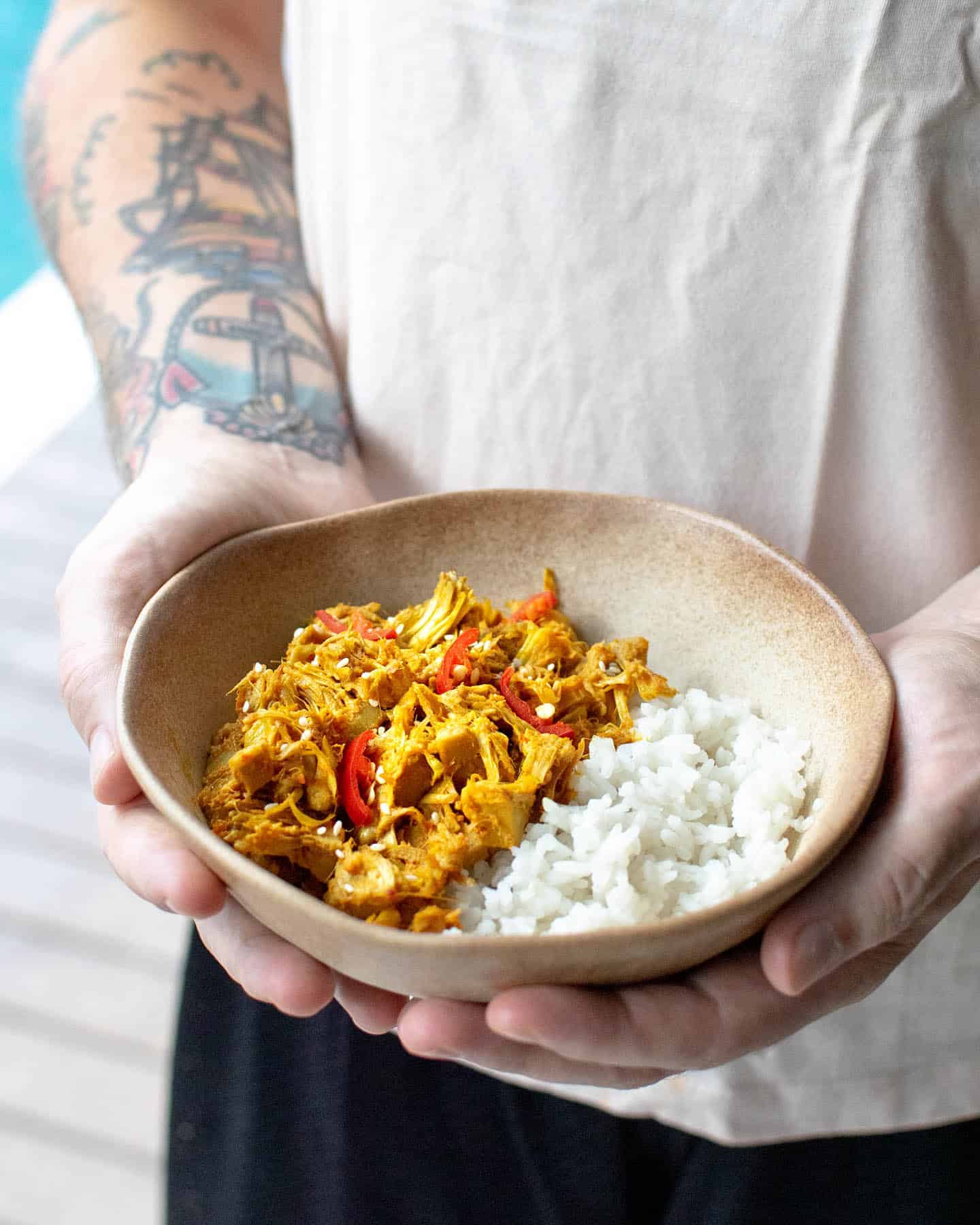 Dan holding a brown bowl of jackfruit rendang and rice that's topped with chopped red chillies and  sesame seeds, decking a a pool can be seen in the background behind Dan's arm