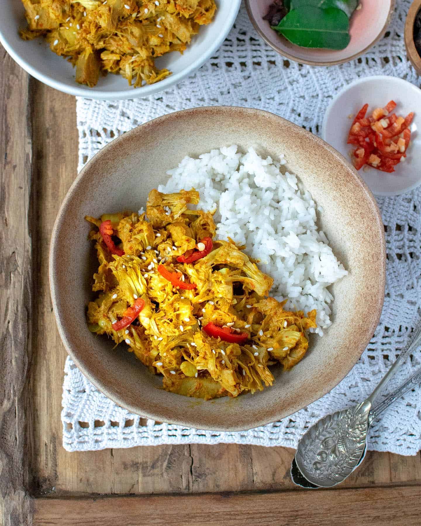 Portrait image, top down view of a brown cowl with Indonesian curry in it and rice, along with small dishes containing red chillies and keffir lime leaves in the background