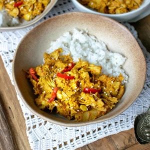 Jackfruit curry in a brown bowl with white rice and fresh red chilli on top, sat on top of a white doily with spoons to the right