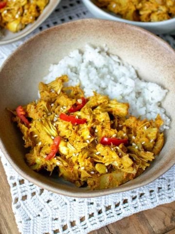 Jackfruit curry in a brown bowl with white rice and fresh red chilli on top, sat on top of a white doily with spoons to the right