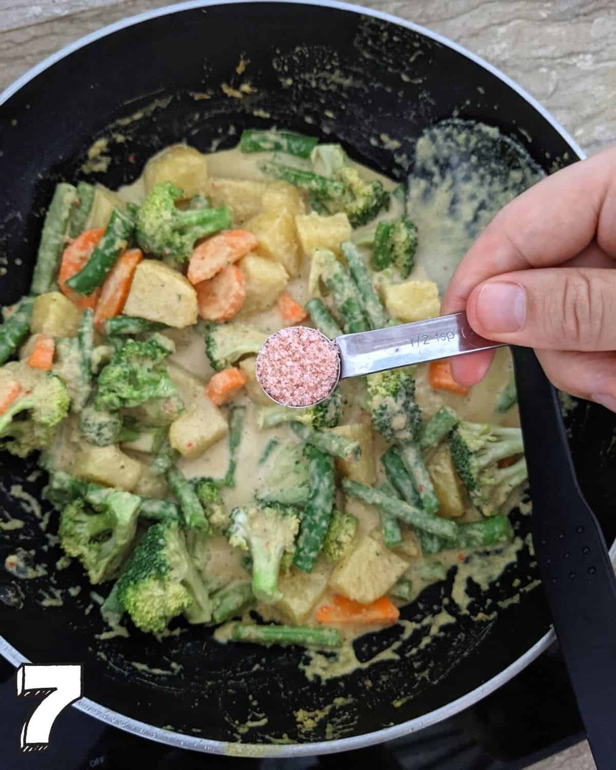 A wok full with sayur kare with a hand holding a spoonful of salt above it