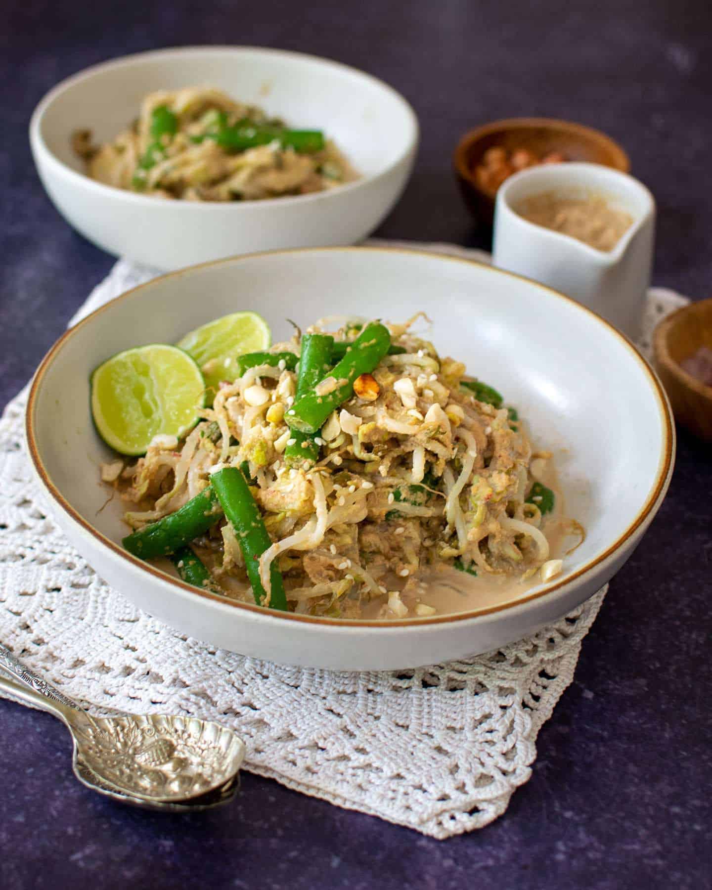 A close up on a white bowl of gado gado salad with spoons to the left hand side resting in front of the bowl and some peanut sauce in the background along with another bowl of salad