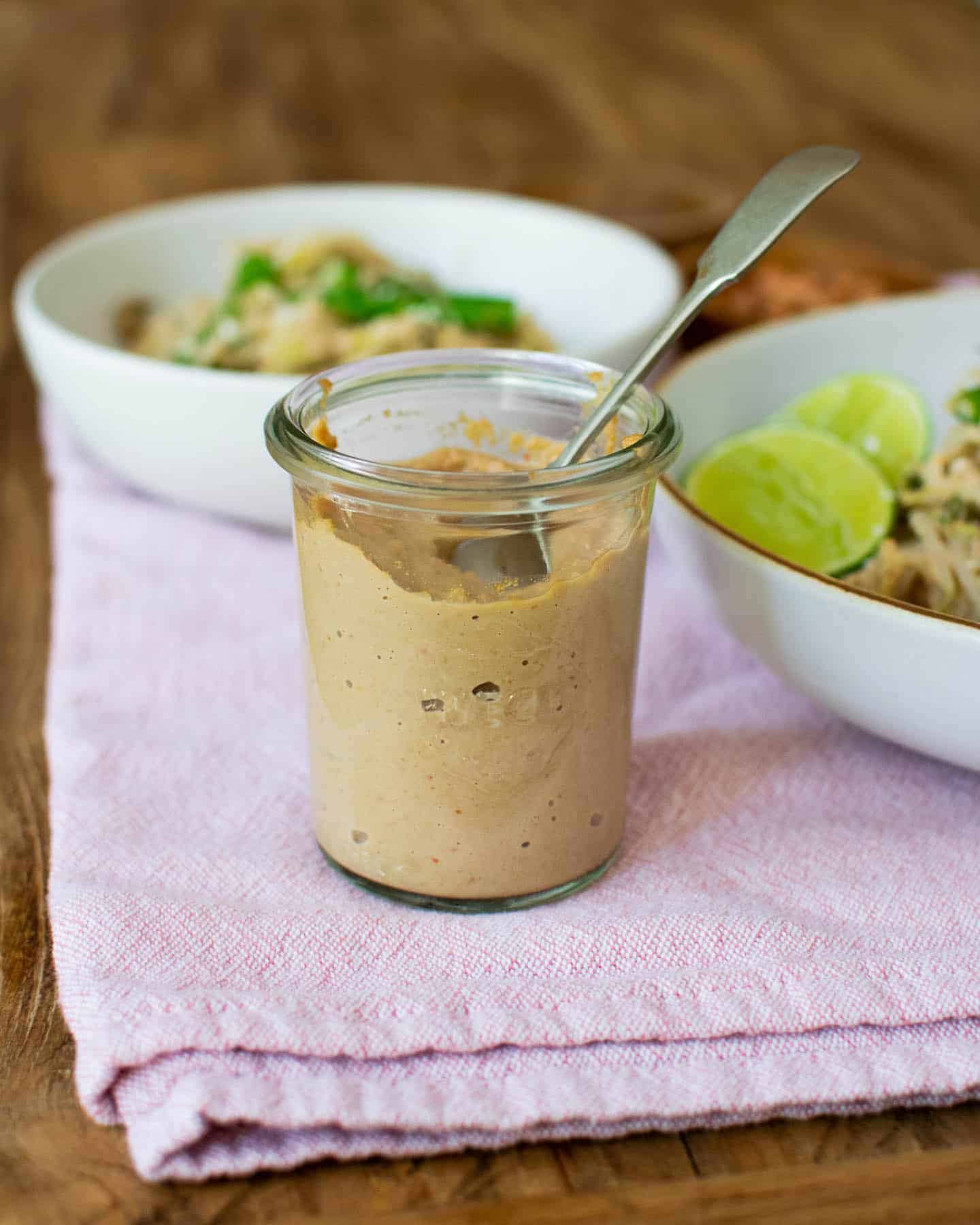 A glass jar filled with peanut sauce with a spoon in it, resting on a pink tea towel with two white bowls to the side and in the background