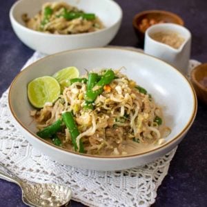 A bowl of vegan gado gado salad topped with green beans, lime halves and peanuts. The angle of the photo is diagonal and there is a ramekin of peanut sauce pointing towards the bowl, and another bowl of salad in the background