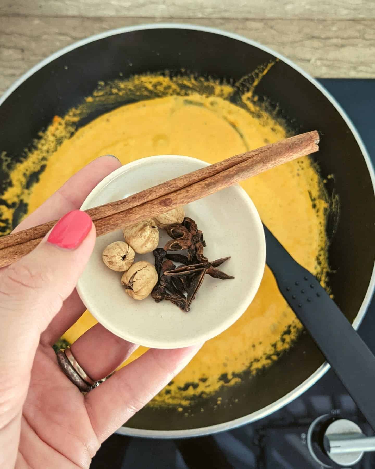 Cinnamon stick, star anise and cardamom pods being held over a wok with sauce in it