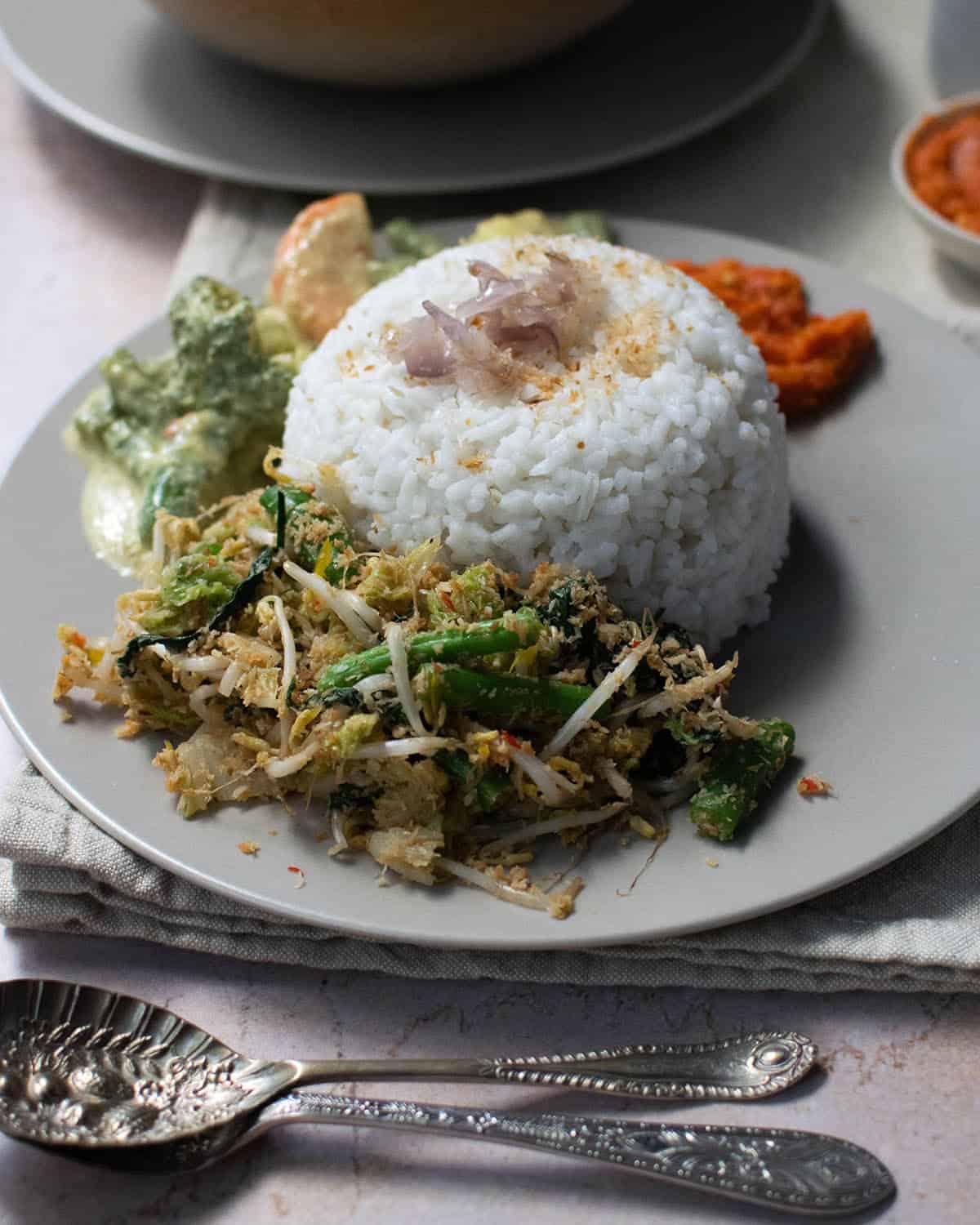 A dome of nasi uduk on a grey plate with veggie dishes around it, and spoons in the foreground