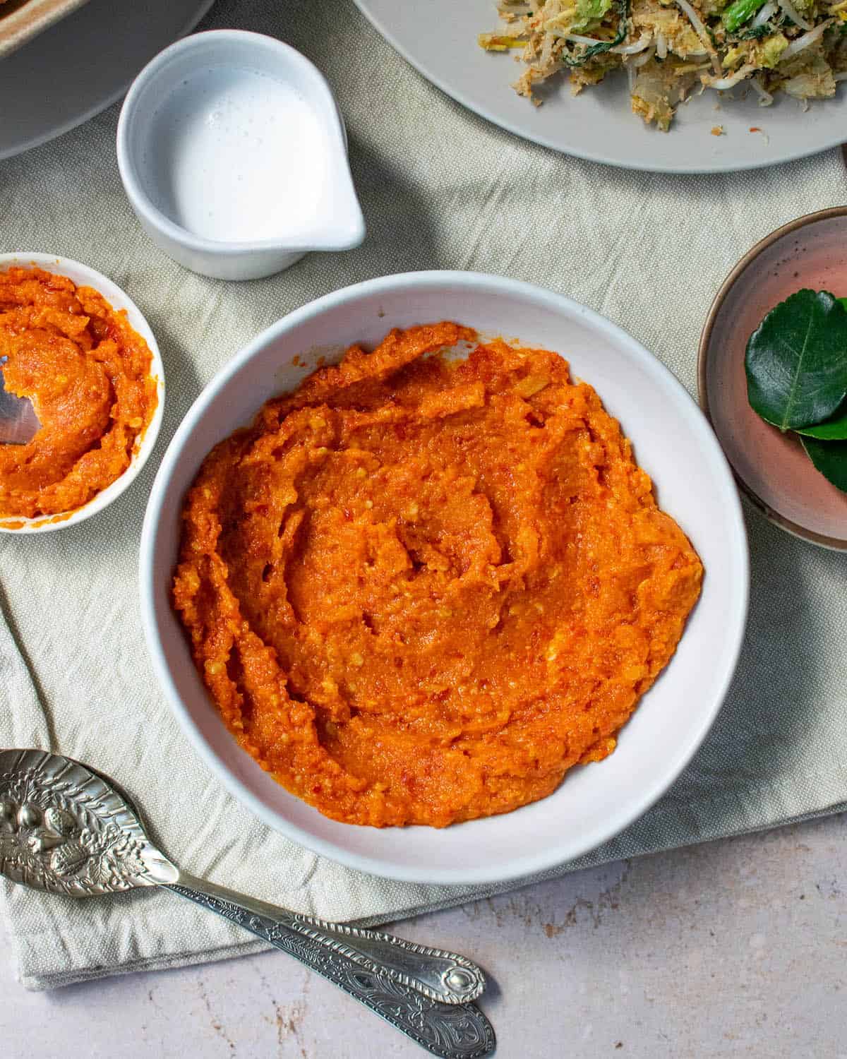 Portrait image showing sambal goreng in a white bowl with spoons, a pot of coconut milk and other plates and dishes surrounding it