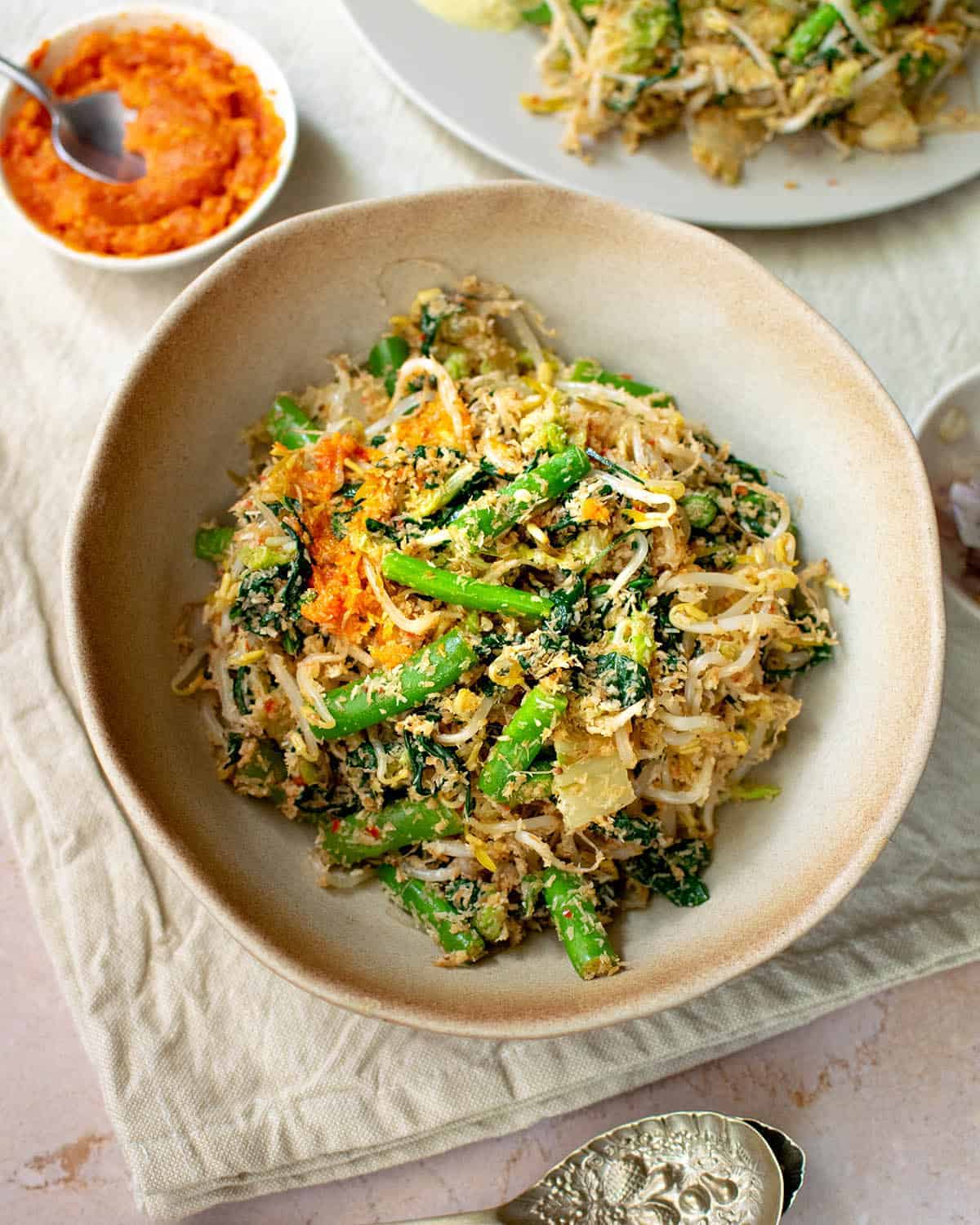 Urab sayur in a brown bowl with some sambal and shallots on top, a spoon in the foreground and sambal and a plate in the background